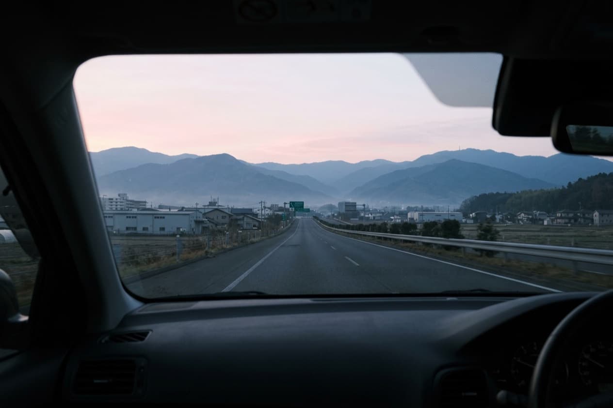 the view forward through a car windscreen at dawn, an open road and blue hills under a pink sky