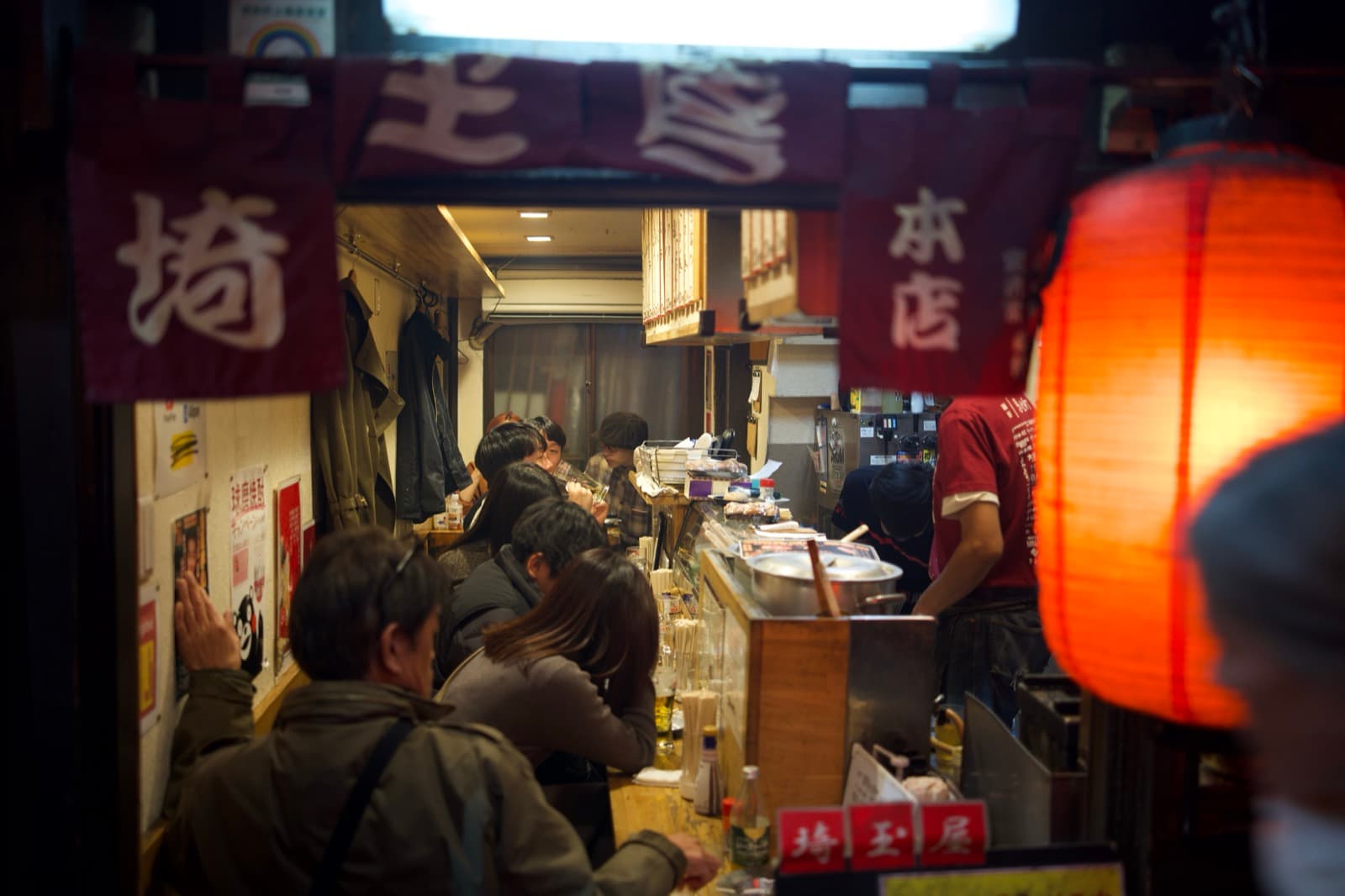 a narrow izakaya counter crowded with drinkers, seen through the noren past a red lantern